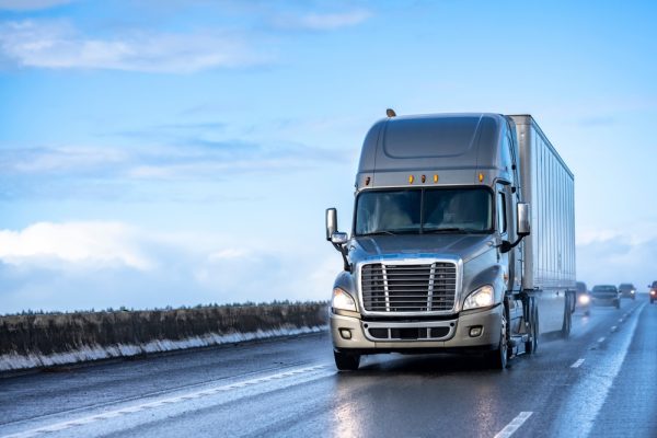 A silver tractor trailer driving over a highway with a reflective hue following a light rain.