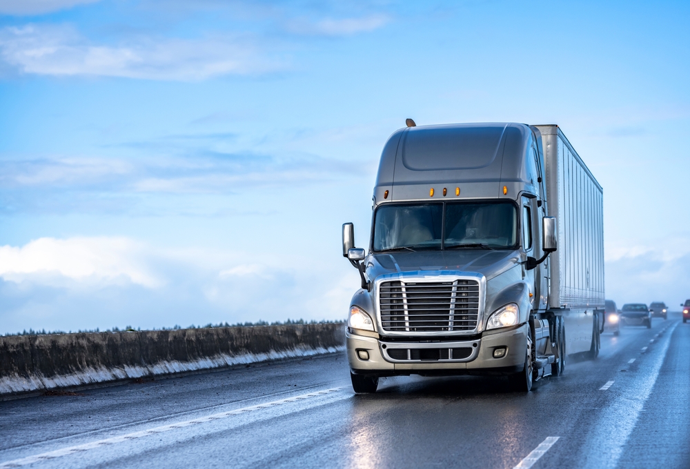 A silver tractor trailer driving over a highway with a reflective hue following a light rain.