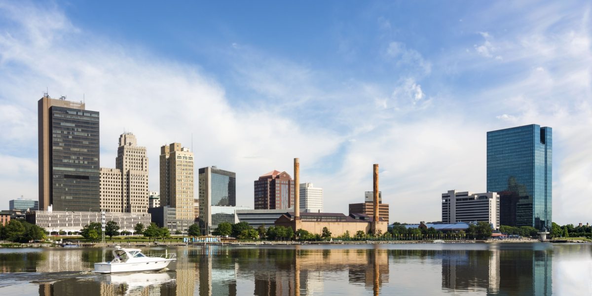 A panoramic view of downtown Toledo Ohio's skyline reflecting into the Maumee river with a power boat cruising by. A beautiful blue sky with white clouds for a backdrop