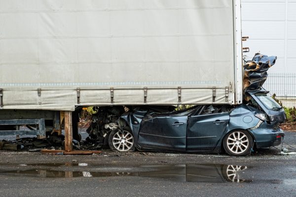 $4 Million Settlement After Passenger Paralyzed in Truck Accident | An image of a light blue sedan crushed beneath a semi-truck's cargo container,.