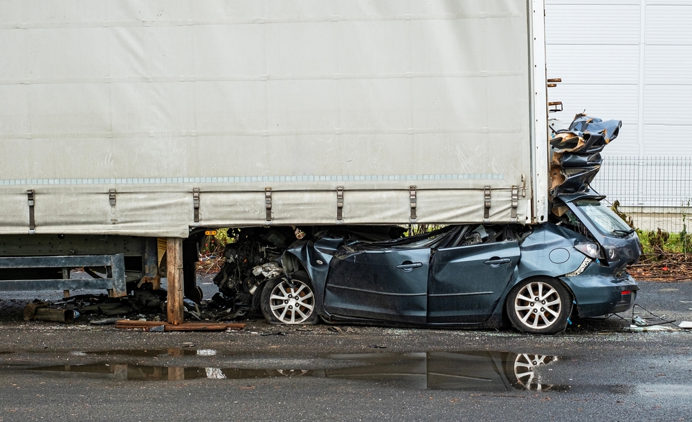 $4 Million Settlement After Passenger Paralyzed in Truck Accident | An image of a light blue sedan crushed beneath a semi-truck's cargo container,.